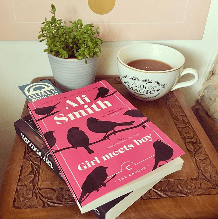 Photograph of a paperback copy of Girl Meets Boy, with a themed bookmark sticking out of the top showing the Queer Book Box logo. It is on top of another book. There is a cup of tea and a small plant in the background. 
