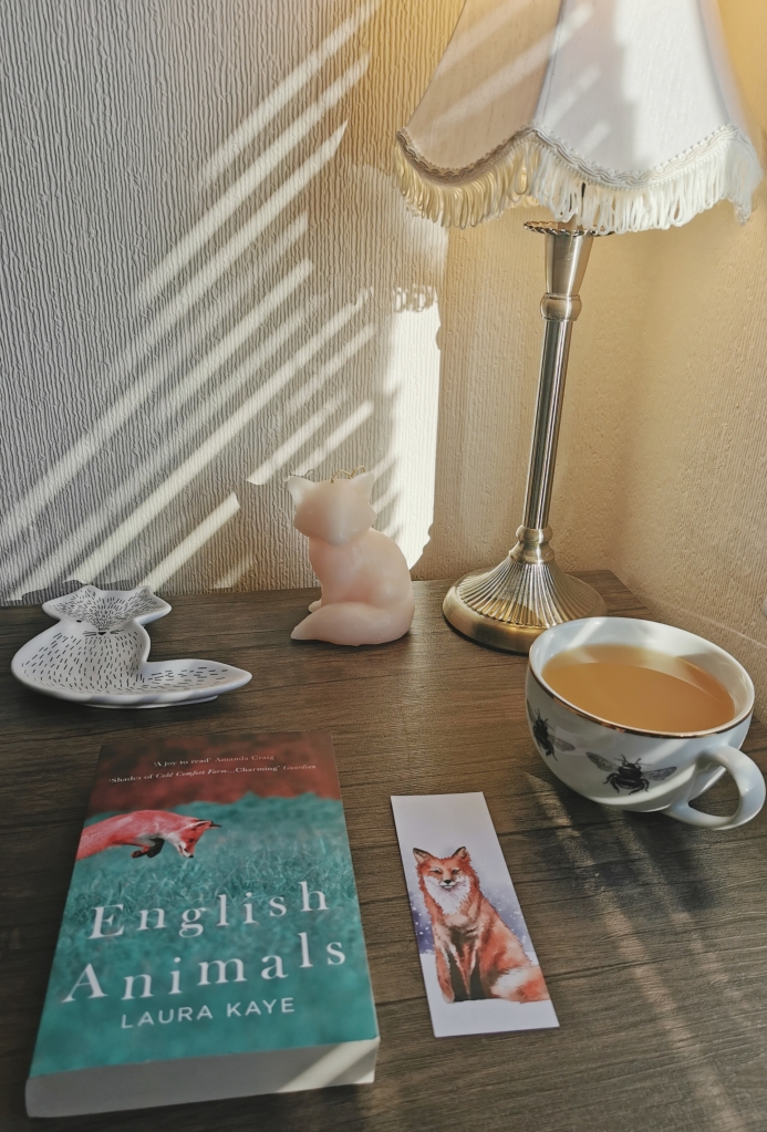 Photograph of a paperback copy of English Animals on a desk, with a themed bookmark. The book and bookmark both have images of foxes on them. In the background is a fox-shaped candle and a fox trinket dish. There is also a cup of tea with a bee design.